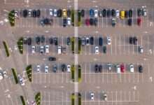 An aerial view of a commercial parking lot. There are many cars parked and they are all of varying colors and designs.