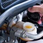 A close-up of a man's hand in the engine bay of a car, lifting the black cap of the brake fluid reservoir.