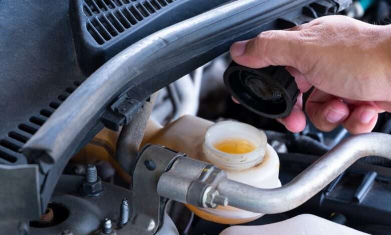 A close-up of a man's hand in the engine bay of a car, lifting the black cap of the brake fluid reservoir.