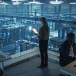 Two cybersecurity colleagues stand in a room overlooking a data center server rack illuminated with multiple fluorescent lights.