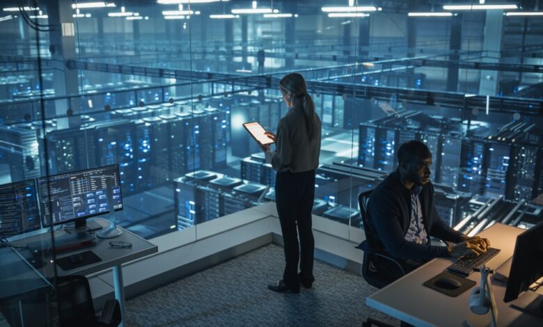 Two cybersecurity colleagues stand in a room overlooking a data center server rack illuminated with multiple fluorescent lights.