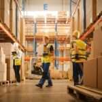 Three warehouse workers in yellow safety vests stand in the middle of a warehouse aisle surrounded by boxes.