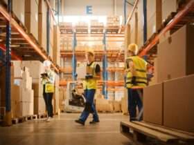 Three warehouse workers in yellow safety vests stand in the middle of a warehouse aisle surrounded by boxes.
