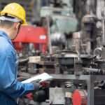 An engineer inspects a machine in a factory. He holds a clipboard and wears a yellow hard hat and blue jumpsuit.