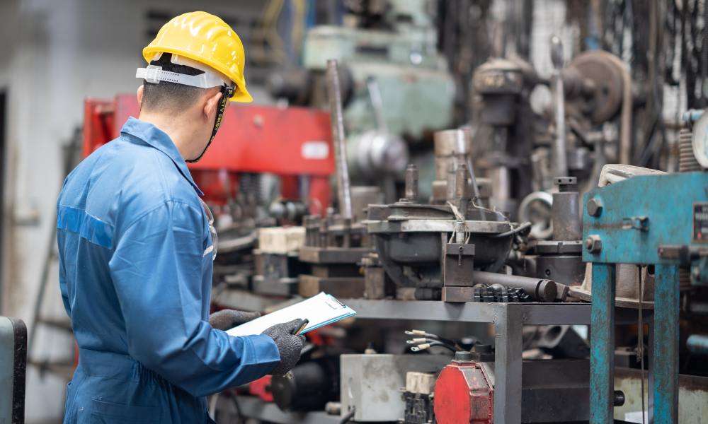 An engineer inspects a machine in a factory. He holds a clipboard and wears a yellow hard hat and blue jumpsuit.