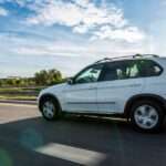 A car is driving down a clear road with the sun casting its shadow on the pavement. The sky above is slightly cloudy.