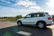 A car is driving down a clear road with the sun casting its shadow on the pavement. The sky above is slightly cloudy.