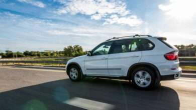 A car is driving down a clear road with the sun casting its shadow on the pavement. The sky above is slightly cloudy.