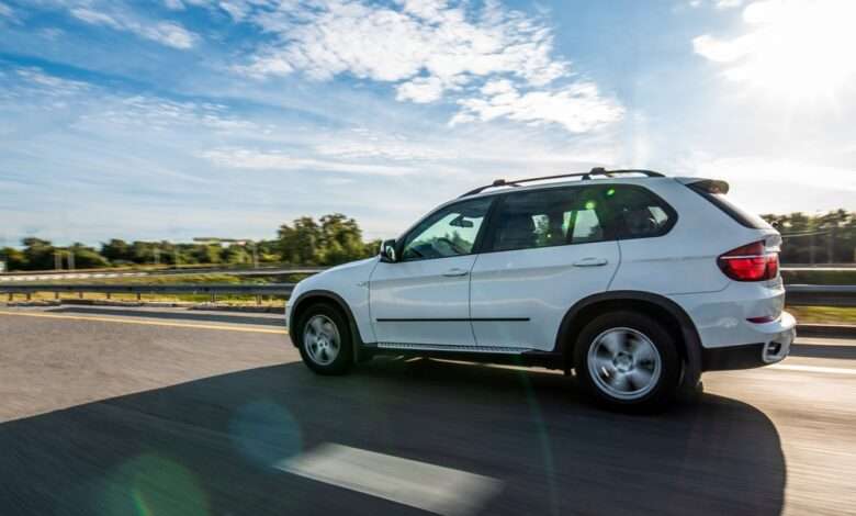 A car is driving down a clear road with the sun casting its shadow on the pavement. The sky above is slightly cloudy.