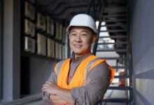 A construction worker wearing a white hard hat and an orange safety vest smiles. He stands in an alleyway.