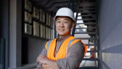 A construction worker wearing a white hard hat and an orange safety vest smiles. He stands in an alleyway.