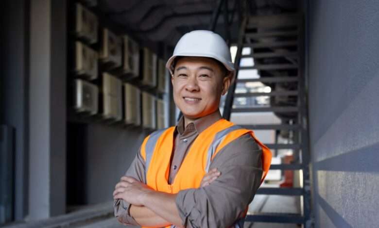 A construction worker wearing a white hard hat and an orange safety vest smiles. He stands in an alleyway.