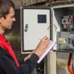 A person looking at an electrical energy meter outside while writing notes in a small purple notebook.