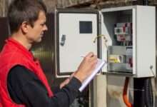 A person looking at an electrical energy meter outside while writing notes in a small purple notebook.