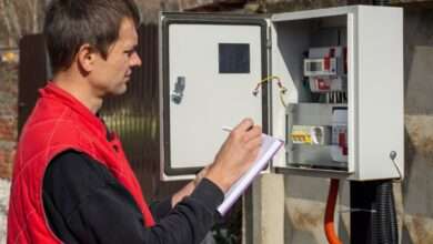 A person looking at an electrical energy meter outside while writing notes in a small purple notebook.