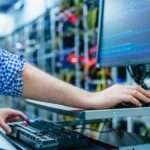A man is sitting in front of a computer screen. In the background are tech components that are blurred.