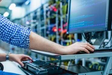 A man is sitting in front of a computer screen. In the background are tech components that are blurred.