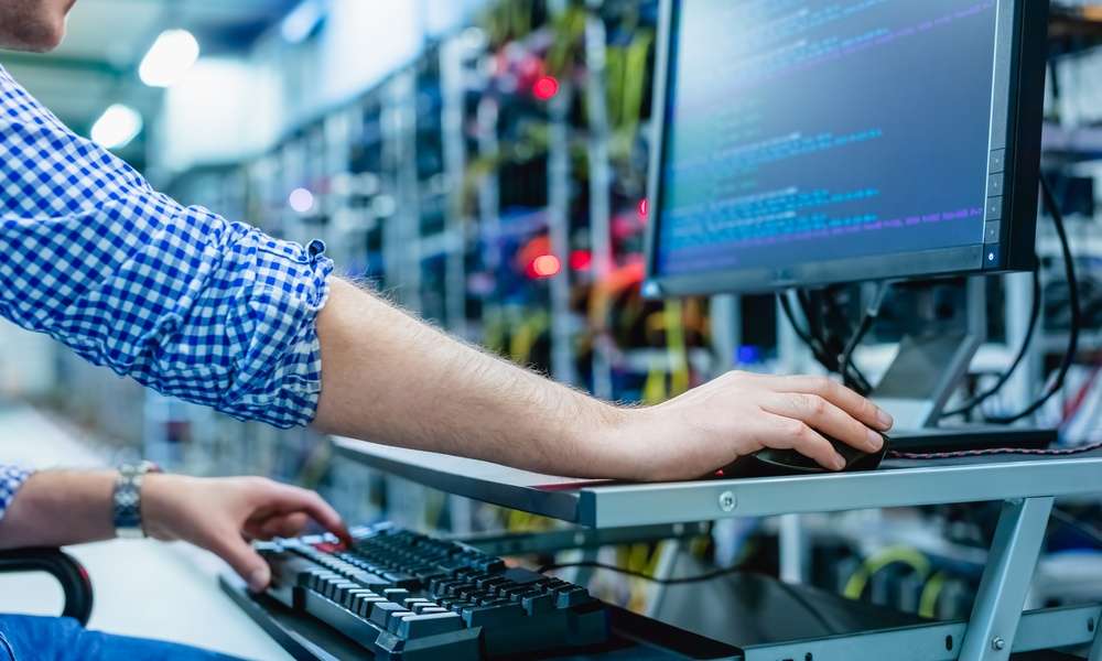 A man is sitting in front of a computer screen. In the background are tech components that are blurred.