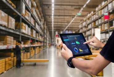 An over-the-shoulder view of a man in a black polo using a smart tablet in the aisle of a large warehouse.