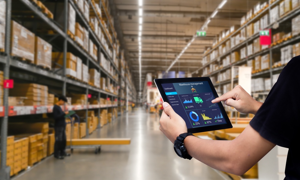 An over-the-shoulder view of a man in a black polo using a smart tablet in the aisle of a large warehouse.