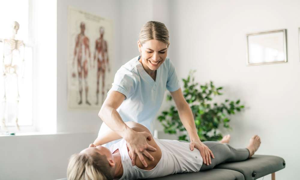 A woman is standing while physically manipulating another woman who is laying down on a table.