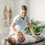 A woman is standing while physically manipulating another woman who is laying down on a table.