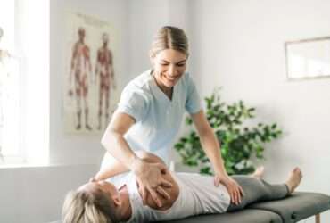 A woman is standing while physically manipulating another woman who is laying down on a table.