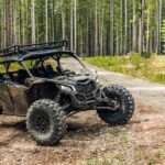 A UTV parked in the woods with tall trees behind it. The UTV is on a dirt path, and its tires are covered in mud.