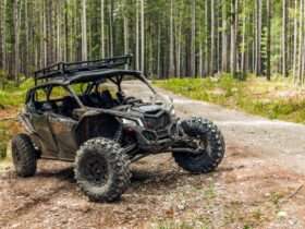 A UTV parked in the woods with tall trees behind it. The UTV is on a dirt path, and its tires are covered in mud.