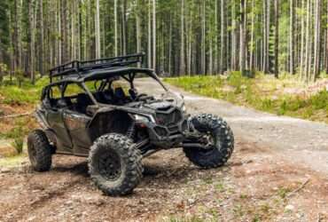 A UTV parked in the woods with tall trees behind it. The UTV is on a dirt path, and its tires are covered in mud.