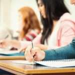 People are sitting at desks; they’re all lined up in a row. The people are writing in notebooks.