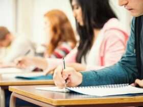 People are sitting at desks; they’re all lined up in a row. The people are writing in notebooks.