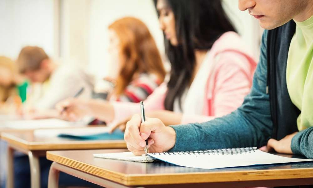 People are sitting at desks; they’re all lined up in a row. The people are writing in notebooks.