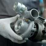 A close-up of a person wearing gray overalls and white gloves. They're holding a new turbocharger for a vehicle.