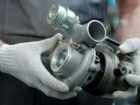 A close-up of a person wearing gray overalls and white gloves. They're holding a new turbocharger for a vehicle.