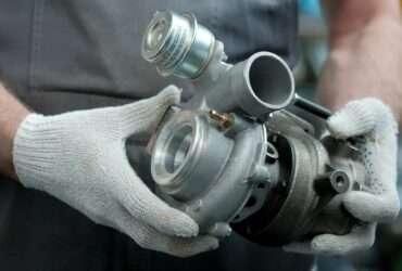A close-up of a person wearing gray overalls and white gloves. They're holding a new turbocharger for a vehicle.