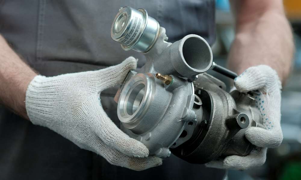 A close-up of a person wearing gray overalls and white gloves. They're holding a new turbocharger for a vehicle.