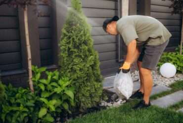 Person wearing gloves picking up yard waste near plants and small tree in landscaped garden beside modern fence.