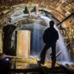 The backside of a person standing in the tunnel of a mine. Water spews out of a pump from the top of the tunnel.