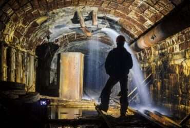 The backside of a person standing in the tunnel of a mine. Water spews out of a pump from the top of the tunnel.