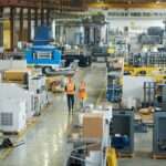 A man and a woman in orange vests are walking in a manufacturing environment. The people and equipment are in a large warehouse.