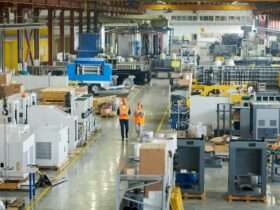 A man and a woman in orange vests are walking in a manufacturing environment. The people and equipment are in a large warehouse.