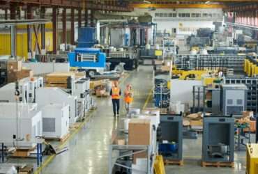 A man and a woman in orange vests are walking in a manufacturing environment. The people and equipment are in a large warehouse.