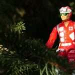 A red action figure dressed as a rescue worker sits among green tree branches, partially surrounded by foliage in natural light.