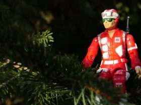 A red action figure dressed as a rescue worker sits among green tree branches, partially surrounded by foliage in natural light.