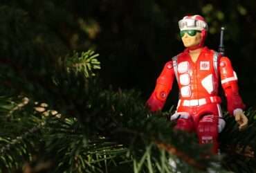 A red action figure dressed as a rescue worker sits among green tree branches, partially surrounded by foliage in natural light.
