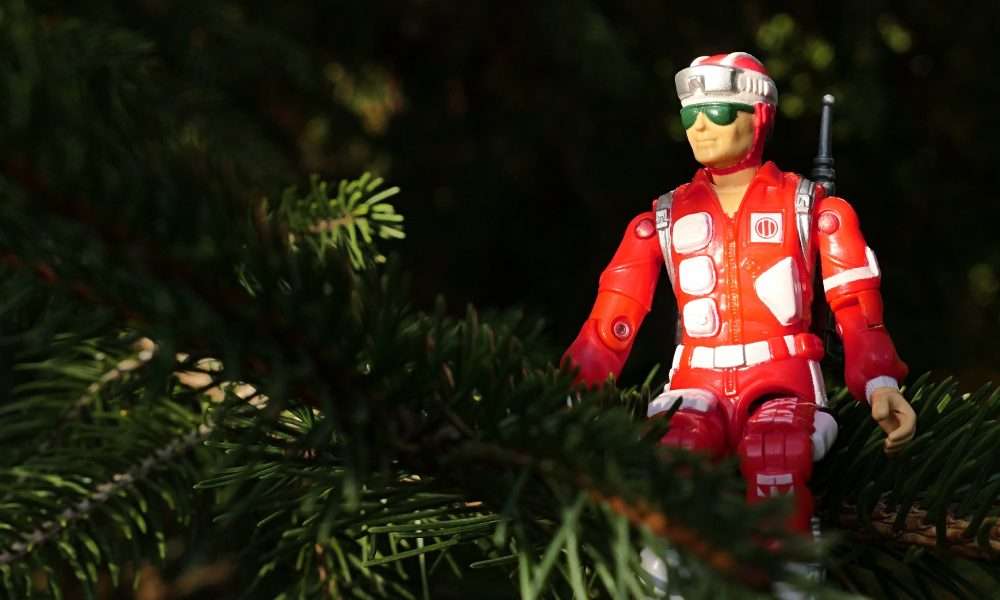 A red action figure dressed as a rescue worker sits among green tree branches, partially surrounded by foliage in natural light.