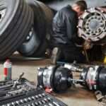 A mechanic works on a truck brake assembly in a workshop, with tools and parts spread across the floor nearby.