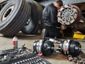 A mechanic works on a truck brake assembly in a workshop, with tools and parts spread across the floor nearby.