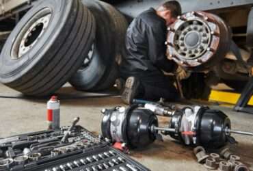 A mechanic works on a truck brake assembly in a workshop, with tools and parts spread across the floor nearby.